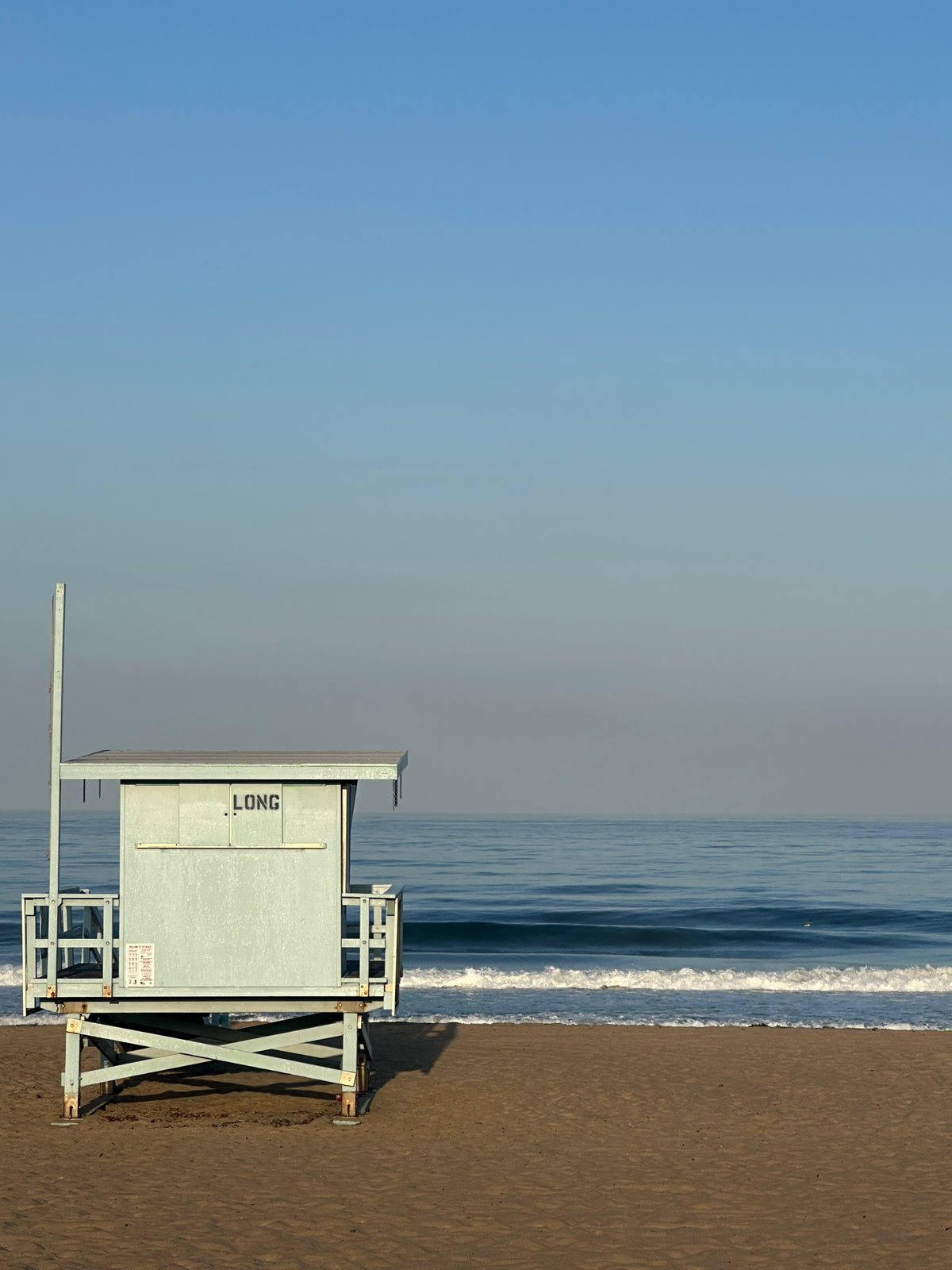 A light blue lifeguard hut stands on a sandy beach with the ocean and a clear blue sky in the background.